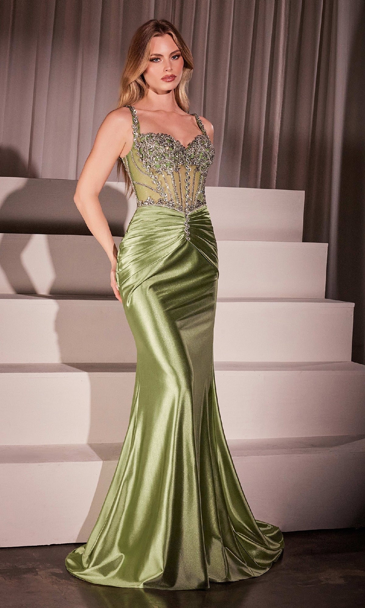 Wearing the Ladivine CD757 Crystal-Bodice Long Prom Dress, a woman stands on a staircase in a fitted, shiny green gown featuring crystal beading and an elegant mermaid silhouette.